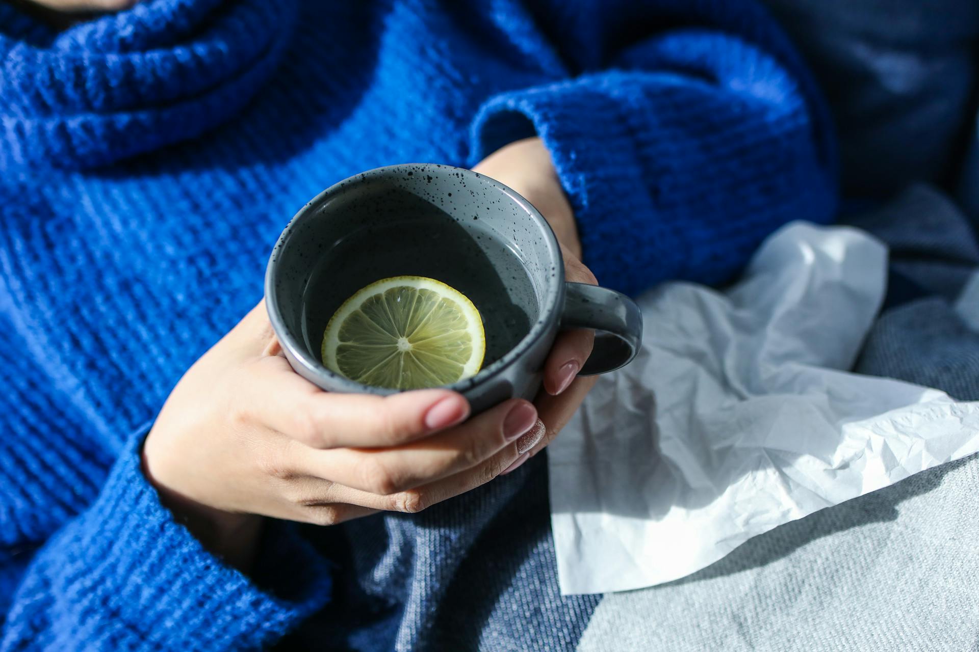Person in a blue jumper holding a hot lemon drink with a tissue nearby, resting at home on a sick day.